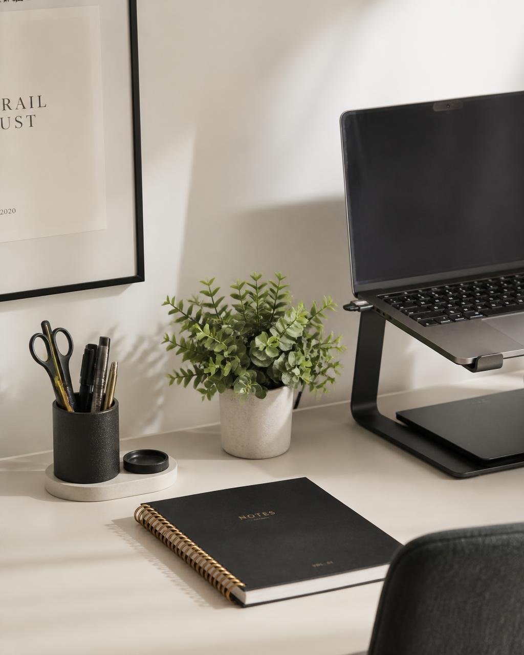 Calm minimal desk corner with a laptop stand, pen cup, notebook and small plant in soft daylight